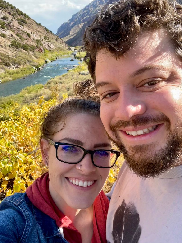 A photo of a man and woman smiling with a river flowing between two mountains and a meadow of yellow flowers in the background.
